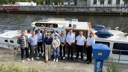 Eine Gruppe von 14 Personen, teils in Uniform, steht auf einem Kai vor einem weißen Boot „MARINE 1“ an einem Fluss mit Graffitimauern.