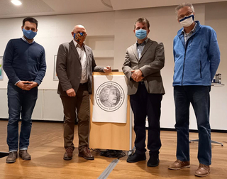 Vier Männer mit Gesichtsmasken stehen nebeneinander vor einem Pult mit Albert-Einstein-Schule-Banner.