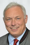 Porträt eines lächelnden älteren Mannes mit grauen Haaren, blauen Augen, in grauer Trachtenjacke und roter Krawatte.