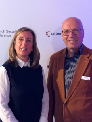 Eine Frau und ein Mann lächeln in die Kamera vor einem Hintergrund der Münchner Sicherheitskonferenz und zeiten.