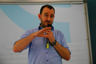 Ein Mann mit Bart und blauem Hemd spricht in ein Mikrofon und gestikuliert während einer Präsentation.