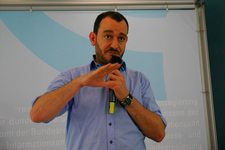 Ein Mann mit Bart und blauem Hemd spricht in ein Mikrofon und gestikuliert während einer Präsentation.
