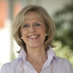 Lächelnde blonde Frau mit Brille, trägt ein helles Hemd mit Kragen und Perlenohrringe.