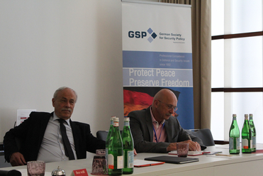 Zwei Männer bei einem Treffen der Deutschen Gesellschaft für Sicherheitspolitik. Ein Banner mit „Protect Peace Preserve Freedom“ ist zu sehen.