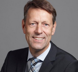 Porträt eines lächelnden Mannes mittleren Alters mit kurzen braunen Haaren, im dunklen Anzug und gemusterter Krawatte.