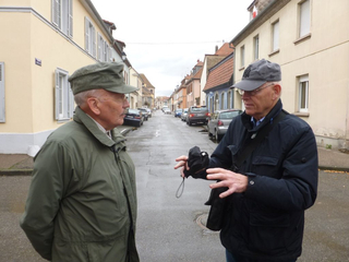 Zwei ältere Männer unterhalten sich auf einer nassen Straße in einer Stadt, einer mit grüner, der andere mit grauer Mütze.
