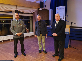 Drei Männer, einer in Uniform, stehen bei einer Veranstaltung der deutschen Reservekräfte. Ein älterer Mann im Anzug ist rechts.