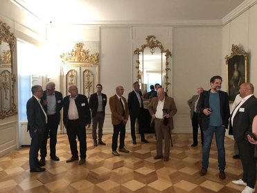 Männer in Anzügen und Jacken stehen in einem prunkvollen Raum mit Goldspiegeln und geometrischem Parkettboden beisammen.