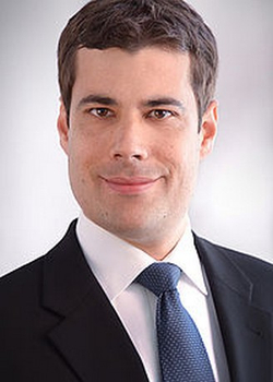 Professionelles Kopfportrait eines lächelnden Mannes mit dunklen Haaren, in dunklem Anzug und blauer Krawatte.