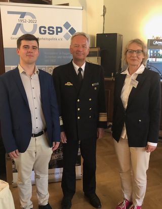 Drei Personen, darunter ein Mann in Marineuniform, posieren vor einem Banner der Gesellschaft für Sicherheitspolitik (GSP).