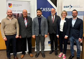 Sechs Personen, vier Männer und zwei Frauen, lächeln vor Veranstaltungsbannern, u.a. Reserve, Cassio, GSP, Uni Bonn.
