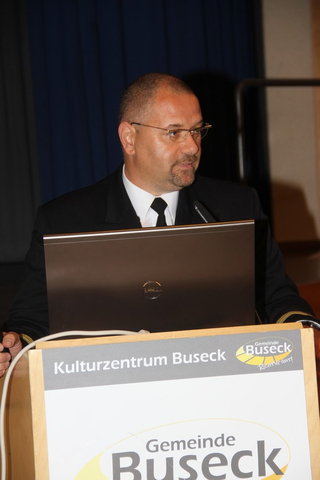 Mann in Uniform spricht hinter Rednerpult mit Laptop und 'Kulturzentrum Buseck'-Logo.