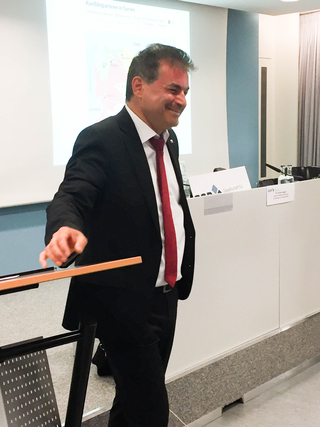 Lächelnder Mann im Anzug und roter Krawatte auf Konferenz, Syrien-Karte auf Leinwand im Hintergrund. Er hält einen Zeigestab.
