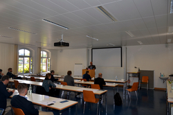 Ein Redner präsentiert in einem Konferenzraum mit Publikum an Tischen, einer Leinwand und hellen Bogenfenstern.