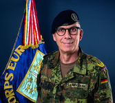 Brigadegeneral Kay Brinkmann Lächelnder deutscher Soldat Brinkmann in Flecktarnuniform und Barett, mit einer 'CORPS EUR'-Flagge im Hintergrund.