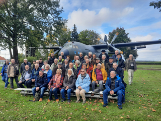 Eine große Gruppe älterer Erwachsener posiert auf einer Wiese vor einem großen Transportflugzeug unter bewölktem Himmel.