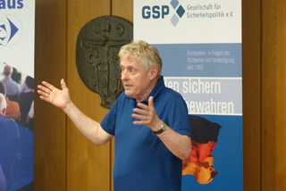 Blonder Mann in blauem Hemd hält einen Vortrag und gestikuliert vor einem GSP-Banner.