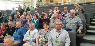 Eine große Gruppe älterer Menschen sitzt in einem Auditorium, viele tragen "Tribüne"-Ausweise, manche schauen sich um oder fotografieren.