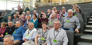 Eine große Gruppe älterer Menschen sitzt in einem Auditorium, viele tragen "Tribüne"-Ausweise, manche schauen sich um oder fotografieren.