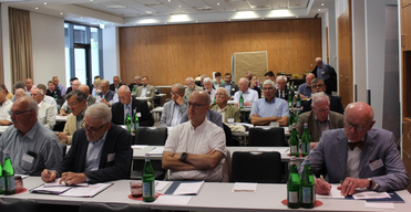 Eine große Gruppe von Teilnehmern, hauptsächlich ältere Männer, sitzt an Tischen in einem Konferenzraum, viele machen Notizen.