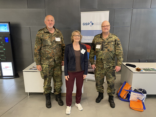 Eine Frau steht zwischen zwei Männern in deutscher Militäruniform bei einer Sicherheitsveranstaltung, mit GSP-Banner im Hintergrund.