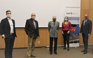 Fünf maskierte Personen, darunter ein Soldat in Uniform, stehen in einem Raum vor einem Banner der Gesellschaft für Sicherheitspolitik.