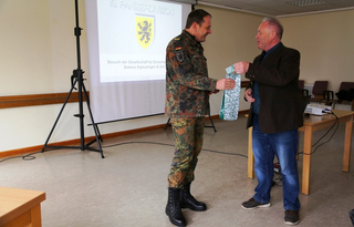 Ein Soldat in Tarnuniform überreicht einem Zivilisten ein Geschenk vor einer Projektionswand mit dem Logo der 10. Panzerdivision.