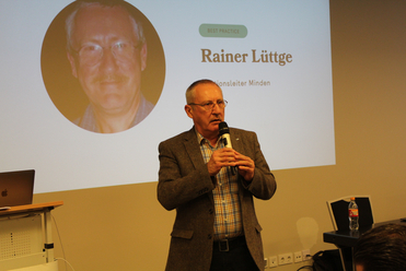 Rainer Lüttge spricht mit Mikrofon vor einer Präsentation, die sein Porträt und seinen Namen zeigt, betitelt 'BEST PRACTICE'.