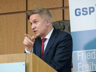 Ein Mann im Anzug und roter Krawatte spricht an einem Holzpult und zeigt gestikulierend. Ein 'GSP'-Banner ist im Hintergrund.