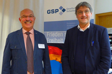 Zwei lächelnde Männer, Josef Schmidhuber und ein Kollege, stehen vor einem GSP-Banner zu Sicherheit, Frieden, Freiheit.