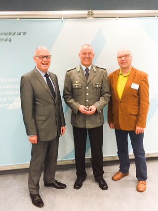 Drei lächelnde Männer, einer in Militäruniform, zwei zivil gekleidet, stehen vor einer blauen Wand mit Text.