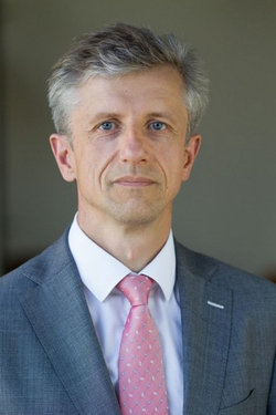 Porträt eines Mannes mit kurzen grauen Haaren und blauen Augen, bekleidet mit grauem Anzug und gemusterter rosa Krawatte.