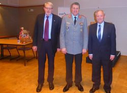 Drei Männer in Anzügen und Uniform, darunter ein Offizier, posieren lächelnd vor einem GSP-Banner bei einer Veranstaltung.