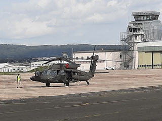 Ein Militärhubschrauber auf einem Flugfeld, daneben eine Person, im Hintergrund ein Kontrollturm und Gebäude.