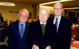 Drei Männer in Anzügen und Krawatten, zwei lächeln, posieren bei einer Indoor-Veranstaltung. Ein Bärtiger, ein älterer Mann, einer mit Brille.