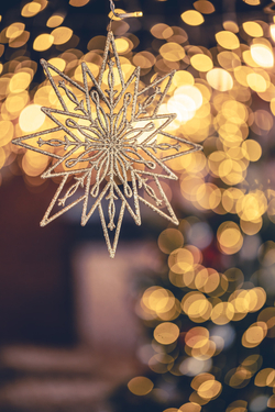 Ein funkelnder goldener Schneeflocken-Anhänger, mit Glitzer verziert, hängt vor einem unscharfen Hintergrund aus warmen Bokeh-Lichtern.