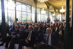 Ein großes Publikum, hauptsächlich Männer in Geschäftskleidung, sitzen in einer großen Halle mit hohen Fenstern und Kronleuchtern bei einer Veranstaltung.