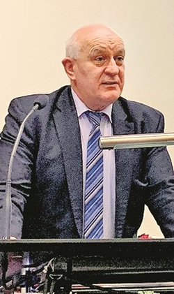 Ein kahler, älterer Mann im dunklen Anzug und blau gestreifter Krawatte spricht an einem Rednerpult mit Mikrofon.