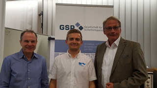 Drei lächelnde Männer vor einem GSP (Gesellschaft für Sicherheitspolitik) Banner. Einer trägt ein MetOp-C Logo-Shirt.