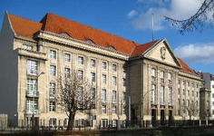 Großes, imposantes Steingebäude mit rotem Ziegeldach und vielen Fenstern, schräg unter blauem Himmel gesehen. Kahle Bäume davor.