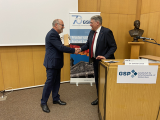 Zwei Männer schütteln sich bei einem GSP-Event die Hände. Dr. Gerhard Conrad steht am Rednerpult, Banner und Büste im Hintergrund.