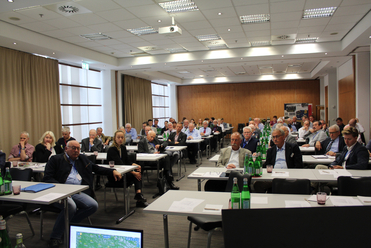 Teilnehmer bei einer Konferenz oder einem Seminar sitzen an Tischen in einem hellen Raum mit Projektor.
