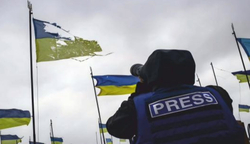 Pressefotograf mit Kamera blickt auf zerfetzte ukrainische Flaggen vor bewölktem Himmel.