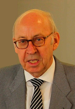 Porträt eines älteren Mannes mit Brille, weißem Hemd, gestreifter Krawatte und grauem Anzug, der zu sprechen scheint.