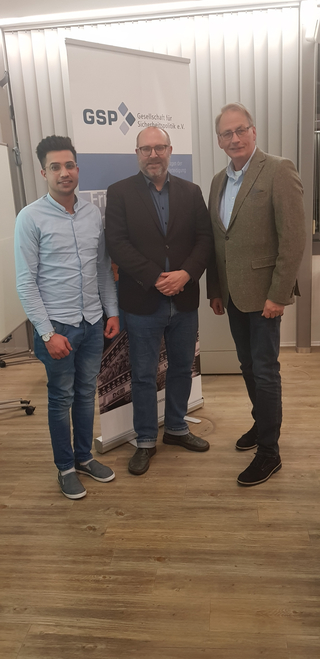 Drei Männer, ein junger und zwei ältere, lächeln vor einem Banner der 'GSP Gesellschaft für Sicherheitspolitik'.