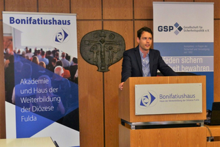 Sprecher bei Bonifatiushaus-Veranstaltung, mit Bannern der 'Akademie und Haus der Weiterbildung der Diözese Fulda' und 'GSP'.