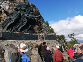 Touristen hören einem Führer vor einem großen bronzenen Kriegerdenkmal mit stürmenden Soldaten an einer felsigen Berglandschaft zu.