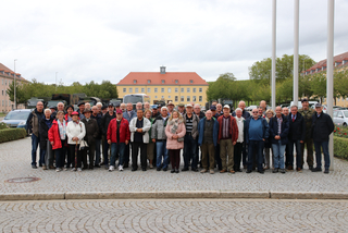 Eine große Gruppe meist älterer Menschen steht auf einem gepflasterten Platz vor Gebäuden und Militärfahrzeugen.