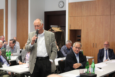 Ein Mann mit Schnurrbart und kariertem Sakko spricht in ein Mikrofon und adressiert eine Gruppe sitzender Männer.