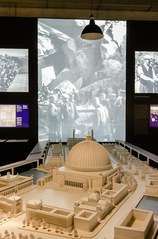 Ein Museumsexponat mit einem großen Modell eines Kuppelgebäudes und Bildschirmen, die historische Fotos von Zwangsarbeit und Menschenmengen zeigen.